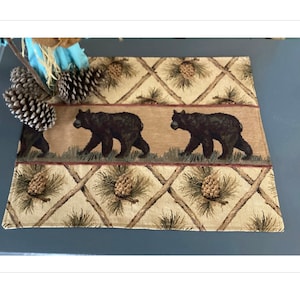 May include: A beige and brown woven placemat with a repeating pattern of pine cones and crossed branches. Two black bears are walking across the placemat.