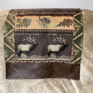 May include: A rectangular furniture cover featuring a central tapestry panel with two elk standing in a field, bordered by pine cones and leaves. The top and bottom sections are made of brown faux leather with a smooth, solid finish.