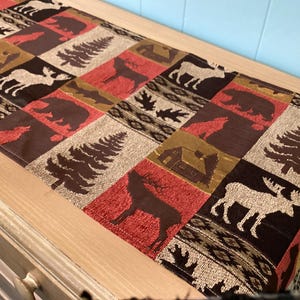 May include: A rectangular table runner with a patchwork design featuring woodland animal silhouettes and pine trees. The color palette includes red, brown, tan, and black. The runner is placed on a light-colored wooden surface.