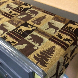 May include: A brown and beige table runner with a woodland theme. The runner features a repeating pattern of moose, bears, deer, and trees.