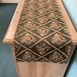 May include: A green table runner with a repeating pattern of pinecones and diagonal lines. The pinecones are tan and brown, set against a dark green background. The runner is displayed on a light-colored wooden table.