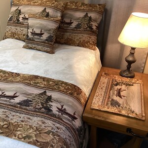 May include: A bed with a cream-colored comforter and matching pillows, adorned with a tapestry featuring a scenic outdoor design. A wooden nightstand holds a lamp and a matching tapestry placemat.