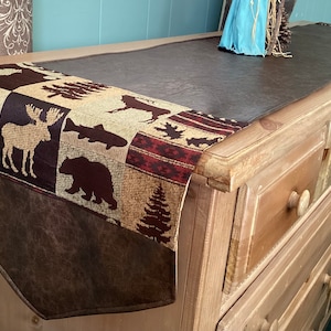 May include: A brown leather table runner with a patterned fabric insert featuring a woodland theme. The fabric insert has images of a cabin, moose, fish, bear, and pine trees. The runner is on top of a light wood dresser with multiple drawers.