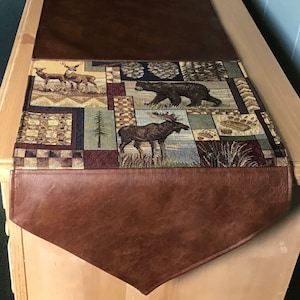 May include: A brown faux leather table runner with a tapestry insert featuring a woodland scene with moose, deer, bears, pine cones, and trees. The tapestry is multi-colored with shades of brown, green, blue, and yellow.
