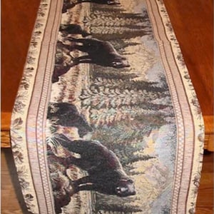 May include: A rectangular table runner featuring a woven tapestry design of black bears walking through a forest of pine trees and mountains. The runner has a decorative border with a repeating leaf pattern in shades of tan, brown, and cream.