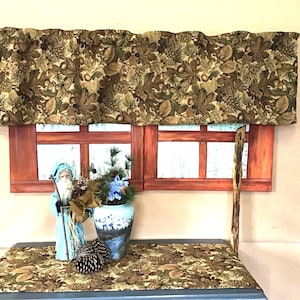 May include: A window valance and matching table runner featuring a brown and green autumn leaf, acorn, and pinecone pattern. A Santa figurine in a blue coat, a blue and white vase with greenery, and two pinecones sit on the table runner.