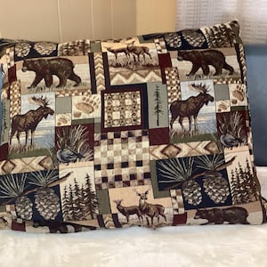 May include: A decorative pillow with a patchwork design featuring bears, moose, deer, pine cones, and trees. The color palette includes shades of brown, green, and burgundy, creating a rustic, outdoorsy theme. The pillow is rectangular.