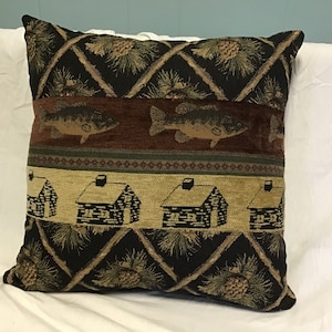 May include: A decorative throw pillow with a black background featuring a repeating pattern of pine cones and branches. The pillow has a brown, green, and beige stripe with images of fish and log cabins.
