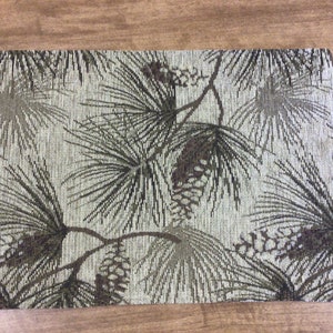 May include: A rectangular placemat with a pine tree and pine cone design. The placemat is a light beige color with dark green and brown pine trees and pine cones.