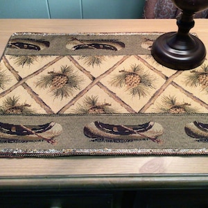 May include: A green and beige table runner with a woven design featuring pine cones, branches, and canoes. The runner has a gold and silver trim.
