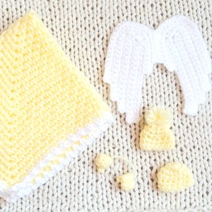 May include: A handmade crochet baby set in soft yellow and white. The set includes a blanket, angel wings, a hat with a pom-pom, and two small accessories. The items are arranged on a textured, off-white surface.