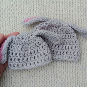 Two gray crochet baby beanie hats are against a cream background. The smaller of the two hats is held by an adult hand. Each hat has two crochet bunny ears attached near the crown.