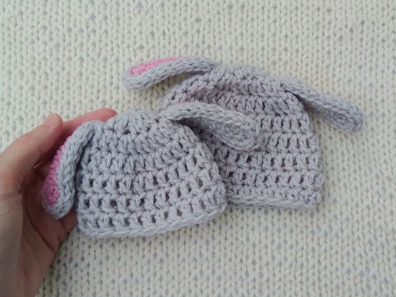 Two gray crochet baby beanie hats are against a cream background. The smaller of the two hats is held by an adult hand. Each hat has two crochet bunny ears attached near the crown.
