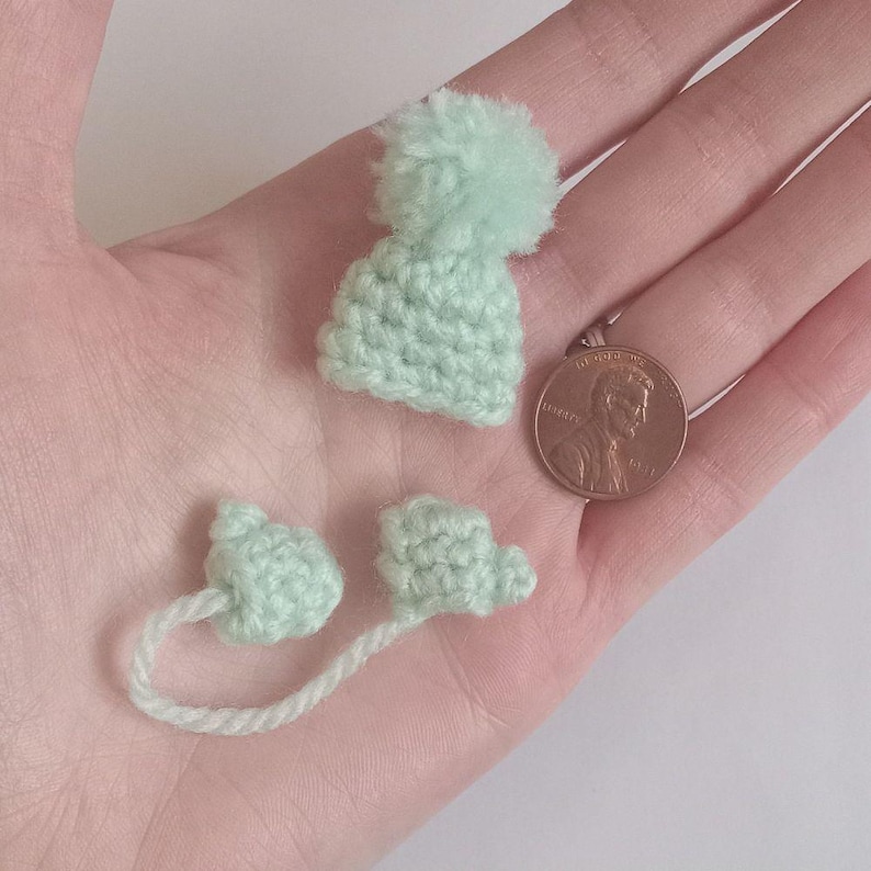 A tiny crochet hat with a pompom rests in an outstretched palm. Below the hat are matching tiny crochet mitts attached with yarn. Next to the st is a US copper penny, to display scale.
