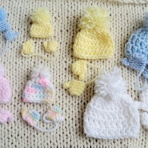Several pastel tiny crochet hats, each with a handmade pom pom rest against a knit cream background. Each pastel hat has a pair of matching mittens corresponding to the size and color of the hat. The smallest set is blue and is for 6 to 8 weeks gestation, while the largest is roughly 20 weeks gestation.