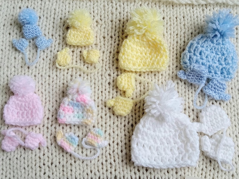 Several pastel tiny crochet hats, each with a handmade pom pom rest against a knit cream background. Each pastel hat has a pair of matching mittens corresponding to the size and color of the hat. The smallest set is blue and is for 6 to 8 weeks gestation, while the largest is roughly 20 weeks gestation.