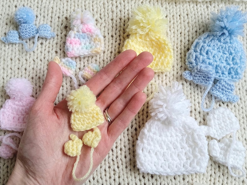 Several pastel tiny crochet hats, each with a handmade pom pom rest against a knit cream background. An outstretched adult hand has a tiny yellow hat in the palm, along with matching yellow mittens that could fit a pea inside. Each pastel hat has a pair of matching mittens corresponding to the size and color of the hat.