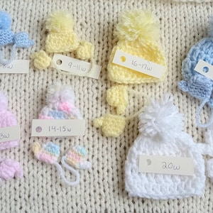 A collection of handmade tiny crochet baby hats in various pastel colors, including pink, yellow, blue, and white, each with a pom-pom. The hats are displayed on a cream-colored knitted surface, with matching miniature mittens underneath the corresponding color and size. Each set has a small card with it indicating what gestational age the set is sized for.