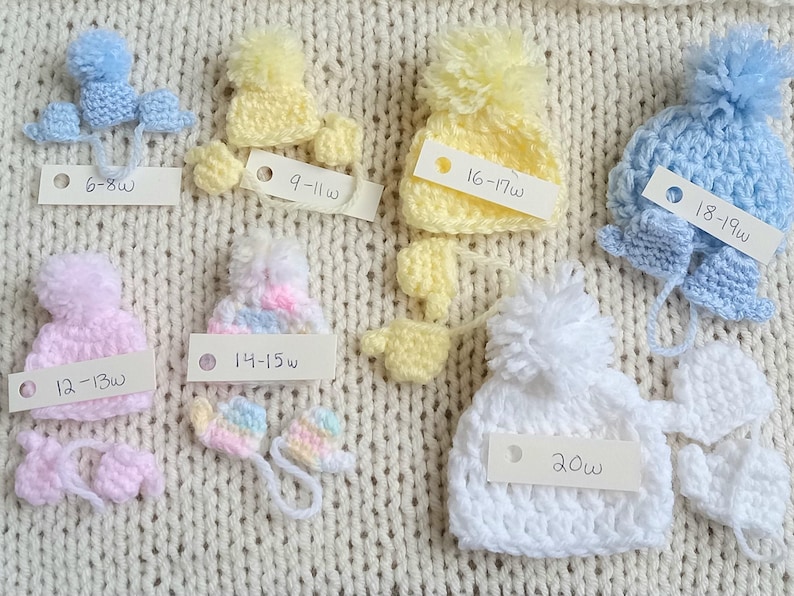 A collection of handmade tiny crochet baby hats in various pastel colors, including pink, yellow, blue, and white, each with a pom-pom. The hats are displayed on a cream-colored knitted surface, with matching miniature mittens underneath the corresponding color and size. Each set has a small card with it indicating what gestational age the set is sized for.