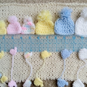 A collection of handmade tiny crochet baby hats in various pastel colors, including pink, yellow, blue, and white, each with a pom-pom. The hats are displayed on a cream-colored knitted surface, with matching miniature mittens underneath the corresponding color and size. There is a ruler in-between the hats and mittens to display scale.