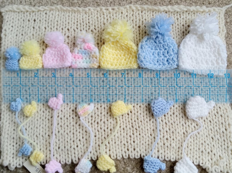 A collection of handmade tiny crochet baby hats in various pastel colors, including pink, yellow, blue, and white, each with a pom-pom. The hats are displayed on a cream-colored knitted surface, with matching miniature mittens underneath the corresponding color and size. There is a ruler in-between the hats and mittens to display scale.