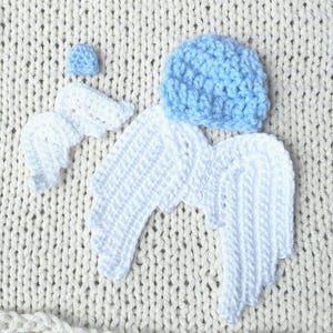 May include: A baby photography set featuring a light blue crocheted beanie, white crocheted angel wings, and a small white crocheted halo. The set is designed for newborn photoshoots and is placed on a cream-colored knitted blanket.