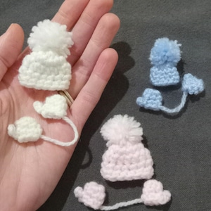 Puede incluir: Tres conjuntos de gorro y mitones de invierno de ganchillo en blanco, azul claro y rosa. Cada gorro tiene un pomp&oacute;n esponjoso y est&aacute; conectado a mitones a juego por un fino hilo de lana. Los art&iacute;culos se muestran sobre un fondo neutro.