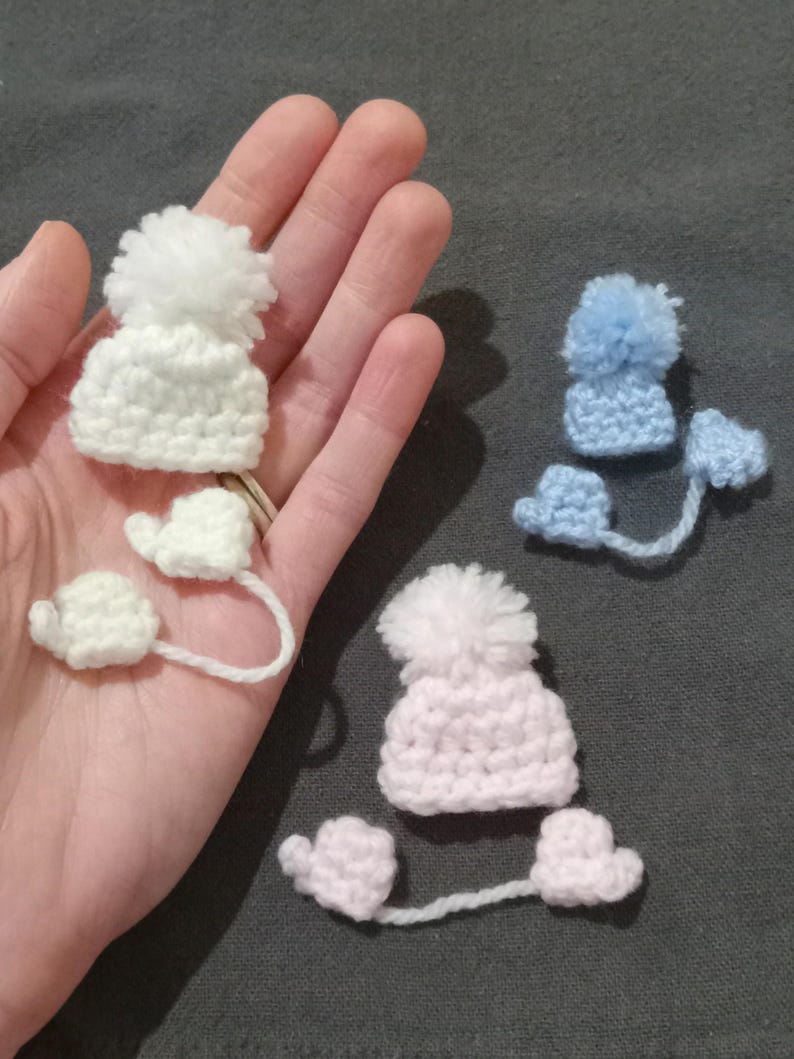Puede incluir: Tres conjuntos de gorro y mitones de invierno de ganchillo en blanco, azul claro y rosa. Cada gorro tiene un pomp&oacute;n esponjoso y est&aacute; conectado a mitones a juego por un fino hilo de lana. Los art&iacute;culos se muestran sobre un fondo neutro.