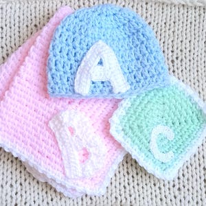 以下が含まれることがあります： 手作りのベビーアイテムのコレクション。白い文字「A」が付いた水色の手編みの帽子、ピンクの毛布、白い文字「B」が付いた白い正方形、白い文字「C」が付いた緑色の正方形。
