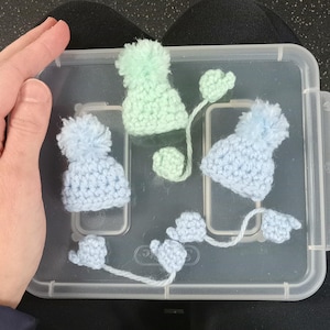 Puede incluir: Una colecci&oacute;n de mini gorros de ganchillo en azul claro y verde menta, cada uno con un pomp&oacute;n esponjoso. Algunos gorros tienen mitones de ganchillo adjuntos. Los art&iacute;culos se muestran dentro de un recipiente de pl&aacute;stico transparente.