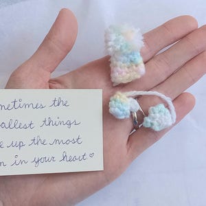 A pair of miniature, pastel-colored crocheted mitts and a matching hat are displayed on an outstretched palm. A handwritten note reads, "Sometimes the smallest things take up the most room in your heart."