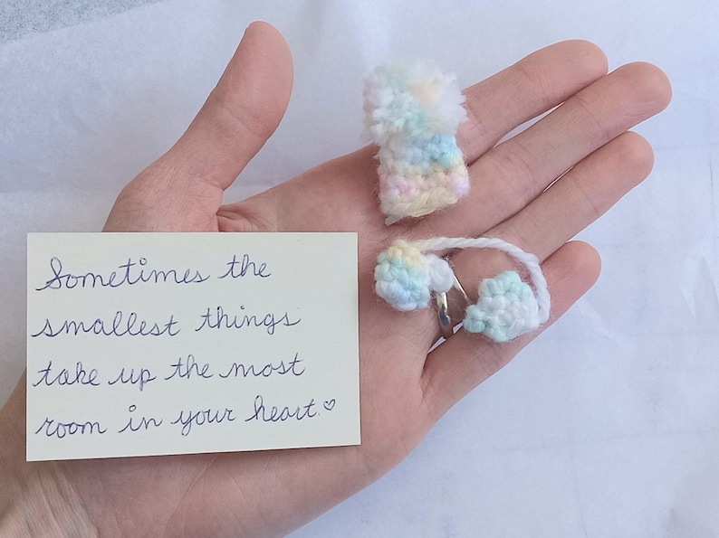 A pair of miniature, pastel-colored crocheted mitts and a matching hat are displayed on an outstretched palm. A handwritten note reads, "Sometimes the smallest things take up the most room in your heart."