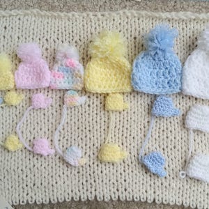 A collection of handmade tiny crochet baby hats in various pastel colors, including pink, yellow, blue, and white, each with a pom-pom. The hats are displayed on a cream-colored knitted surface, with matching miniature mittens underneath the corresponding color and size.