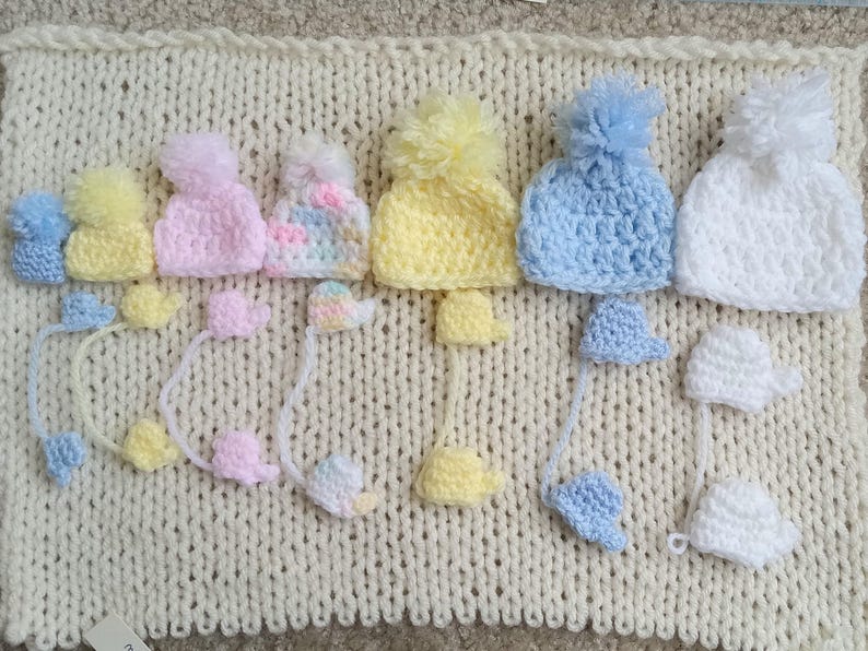 A collection of handmade tiny crochet baby hats in various pastel colors, including pink, yellow, blue, and white, each with a pom-pom. The hats are displayed on a cream-colored knitted surface, with matching miniature mittens underneath the corresponding color and size.