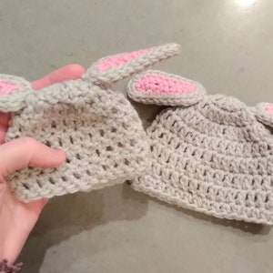 Two gray crochet baby hats are shown against a darker gray background. The smaller of the two hats is held by a hand. Each hat is small and has two crochet bunny ears attached at the top.