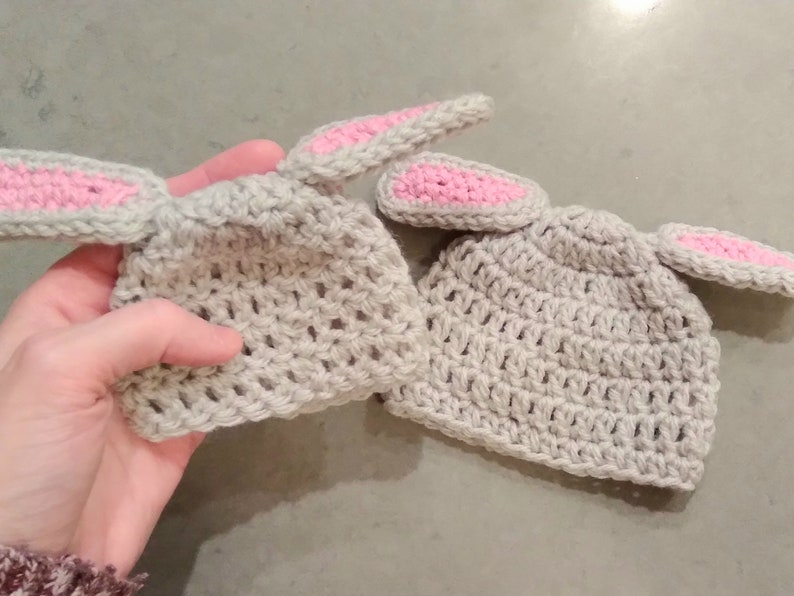 Two gray crochet baby hats are shown against a darker gray background. The smaller of the two hats is held by a hand. Each hat is small and has two crochet bunny ears attached at the top.