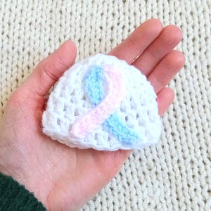 Könnte beinhalten: Ein kleiner, weiß gehäkelter Babyhut mit einem rosa und blauen Banddesign. Der Hut wird in einer Hand gehalten, und der Hintergrund ist ein cremefarbener Strickstoff. Der Hut ist ein handgefertigter Artikel.