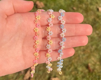 May include: Three beaded flower bracelets in pink, yellow, and blue hues. Each bracelet features small, colorful flower beads with gold-colored centers. The bracelets are held in a hand, showcasing their delicate design and vibrant colors. The bracelets are a charming accessory.