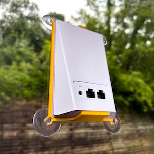 Puede incluir: Un dispositivo electrónico blanco y amarillo con dos puertos negros y un pequeño puerto redondo. El dispositivo está montado en una ventana con ventosas. El fondo es una vista borrosa de árboles verdes.