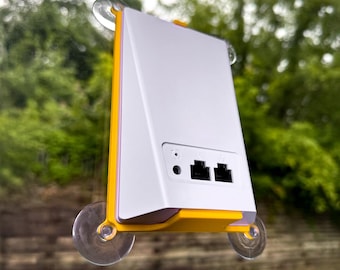 May include: A white and yellow electronic device with two black ports and a small round port. The device is mounted on a window with suction cups. The background is a blurred view of green trees.