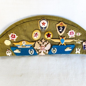 May include: A green military cap with various metal badges and insignia, including a red star, a tank, and a winged emblem. The cap has a number "2" on one of the badges.