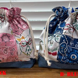 Peut inclure: Deux sacs à cordon avec des bases en denim. L'un est rose avec un imprimé cachemire et chat, l'autre est bleu avec un imprimé cachemire et chat. Les deux sacs ont une fermeture à cordon et un cordon blanc. Le texte "A-PINK" et "B-BLUE" est visible.