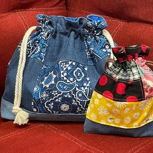 May include: A clear plastic water bottle next to a large denim drawstring pouch with blue and white paisley fabric panels and thick white rope cords, and a smaller drawstring pouch featuring black, red, yellow, and floral patterned fabric panels.