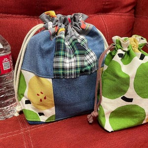Peut inclure: Deux sacs à cordon en tissu. Le plus grand sac présente un motif patchwork composé de denim bleu, de carreaux verts et blancs, et d'un imprimé de fruits jaunes et verts. Le plus petit sac a un motif de fruits verts et jaunes avec des cordons marron.