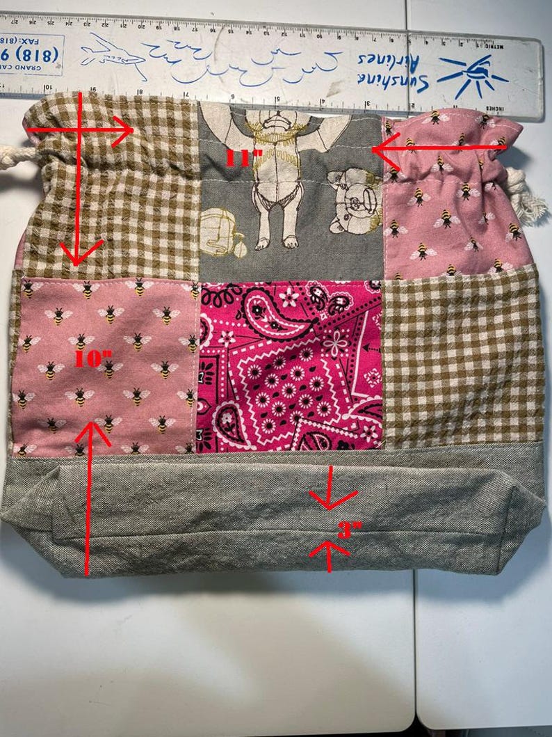 May include: A patchwork drawstring bag with various fabrics. The bag includes pink fabric with bees, brown gingham, pink paisley, and grey fabric. The bag is approx. 25 cm tall, 28 cm wide, and 7.6 cm deep.