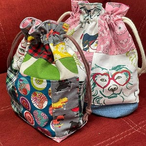 May include: Two fabric drawstring pouches featuring patchwork designs. The left pouch displays a mix of green, blue, and gray panels with patterns of circles, plaid, and small dogs on skateboards. The right pouch features pink, white, and blue denim fabric with a graphic illustration of a dog wearing red heart-shaped glasses.