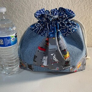 Pode incluir: Uma bolsa de ganga com cord&atilde;o com uma parte superior estampada em azul e branco e um painel cinzento com c&atilde;es em skates. Uma garrafa de &aacute;gua transparente com tampa branca est&aacute; ao lado da bolsa.