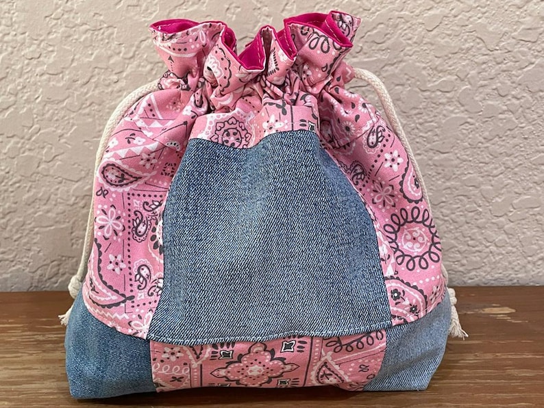 K&ouml;nnte beinhalten: Ein Kordelzugbeutel mit einem Jeans-Mittelteil und rosa Paisley-Stoff oben und unten. Der rosa Stoff hat ein wei&szlig;es Paisley-Design. Die Tasche hat einen wei&szlig;en Kordelzugverschluss.