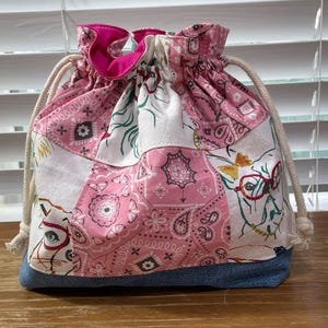 May include: A drawstring bag with a patchwork design. The bag features a mix of pink paisley, white, and denim fabrics. The top is gathered with a pink lining and a drawstring closure. The bag has a whimsical design with a dog wearing glasses.