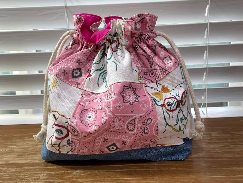 May include: A drawstring bag with a patchwork design. The bag features a mix of pink paisley, white, and denim fabrics. The top is gathered with a pink lining and a drawstring closure. The bag has a whimsical design with a dog wearing glasses.